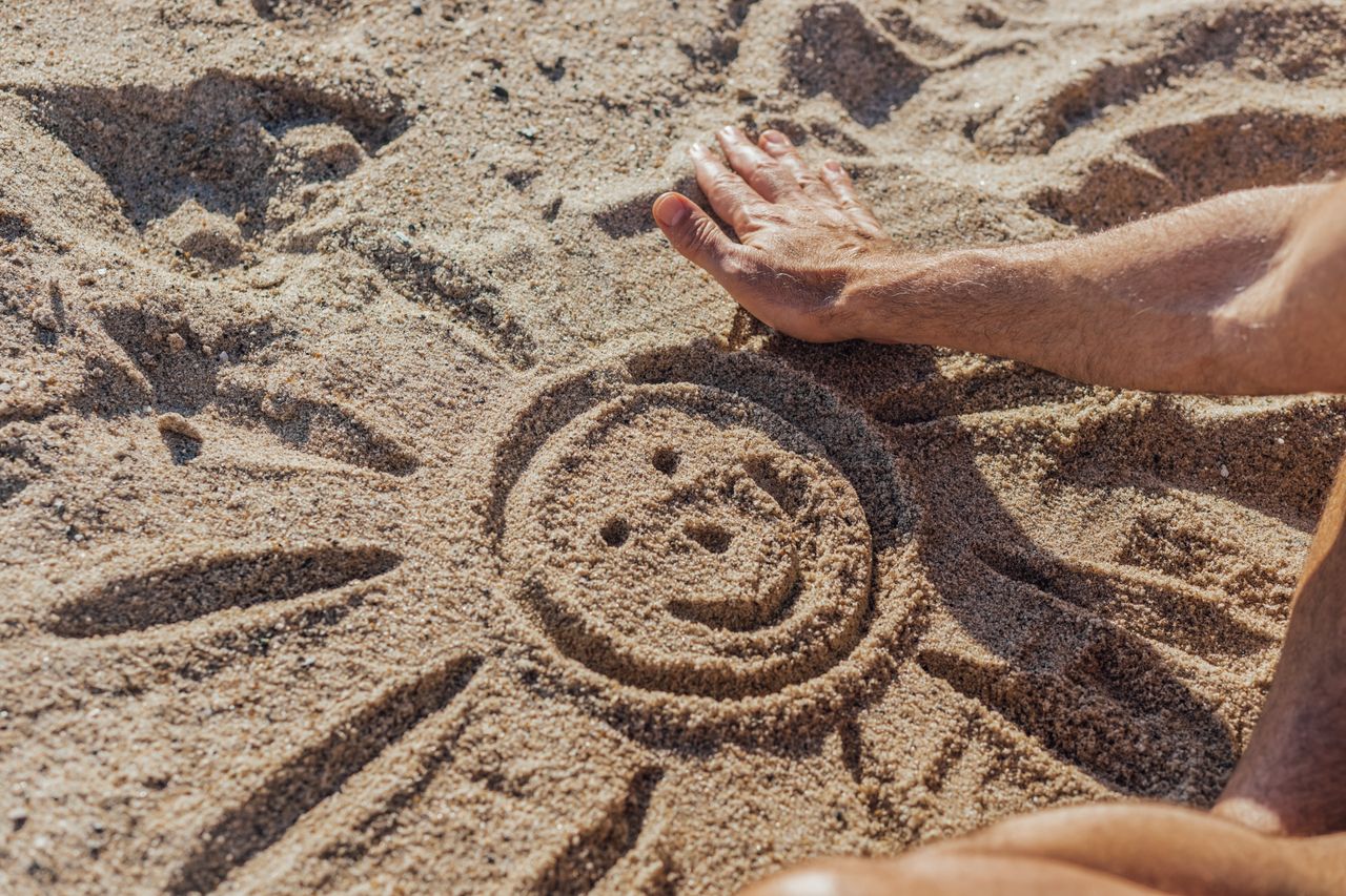 Person creating a cheerful face in the sand on a sunny beach day.